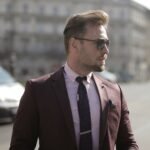 Businessman in sunglasses wearing maroon suit and tie walks confidently in urban setting.